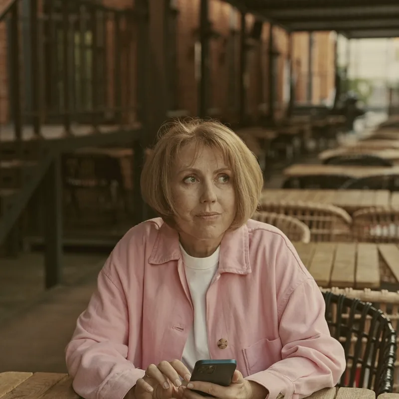 A woman sitting alone with her phone at a long row of empty café tables.