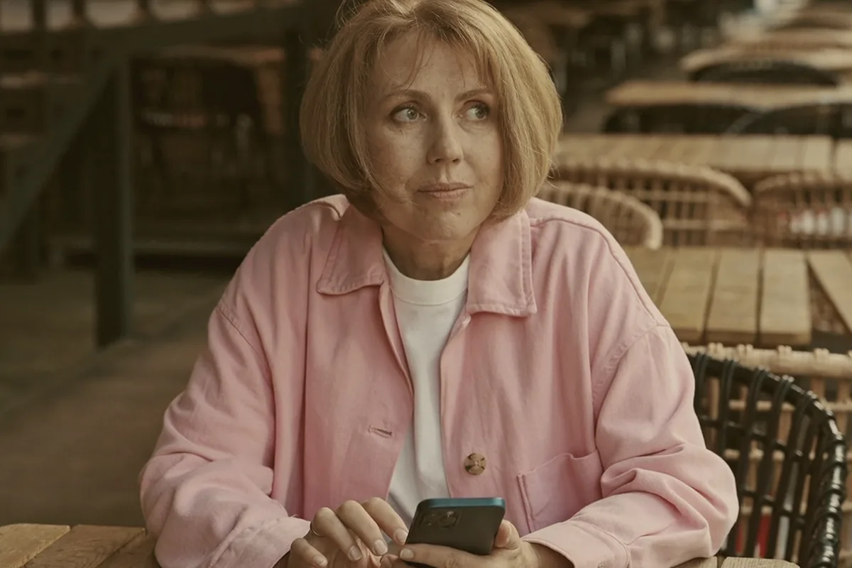 A woman sitting alone with her phone at a long row of empty café tables.